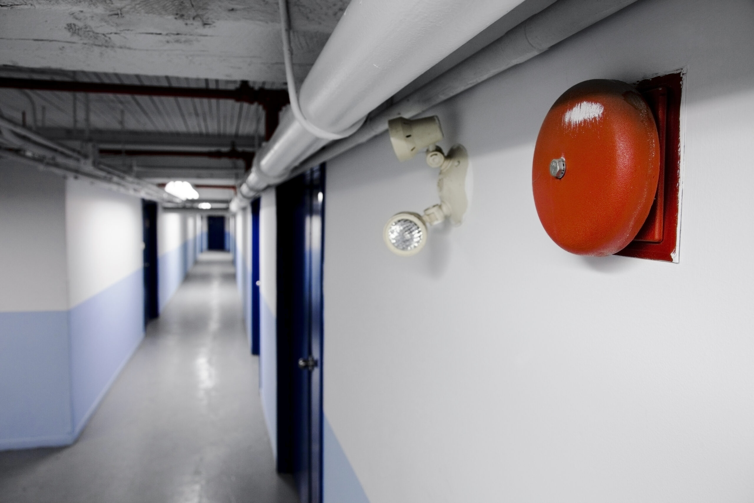 A view of a hallway focussing upon Fire Alarm Bell red in color alongside with the emergency light bulbs installed.